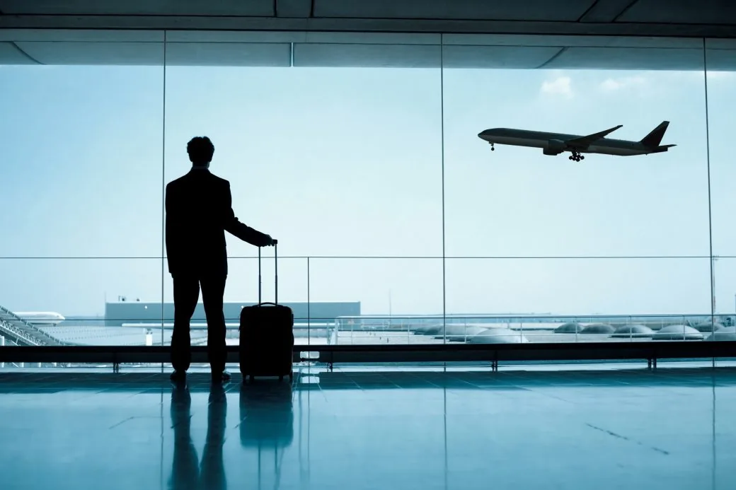 A traveler with a case at a departure lounge window, watching an aircraft take off.