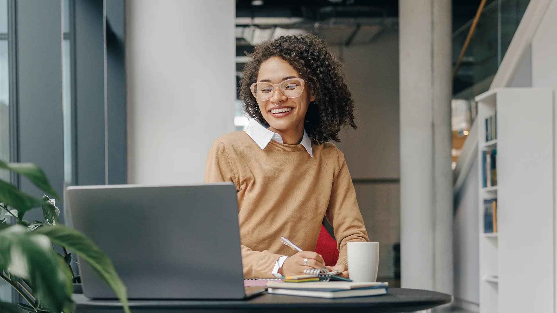 A woman smiling at her laptop at a table in a bright office.